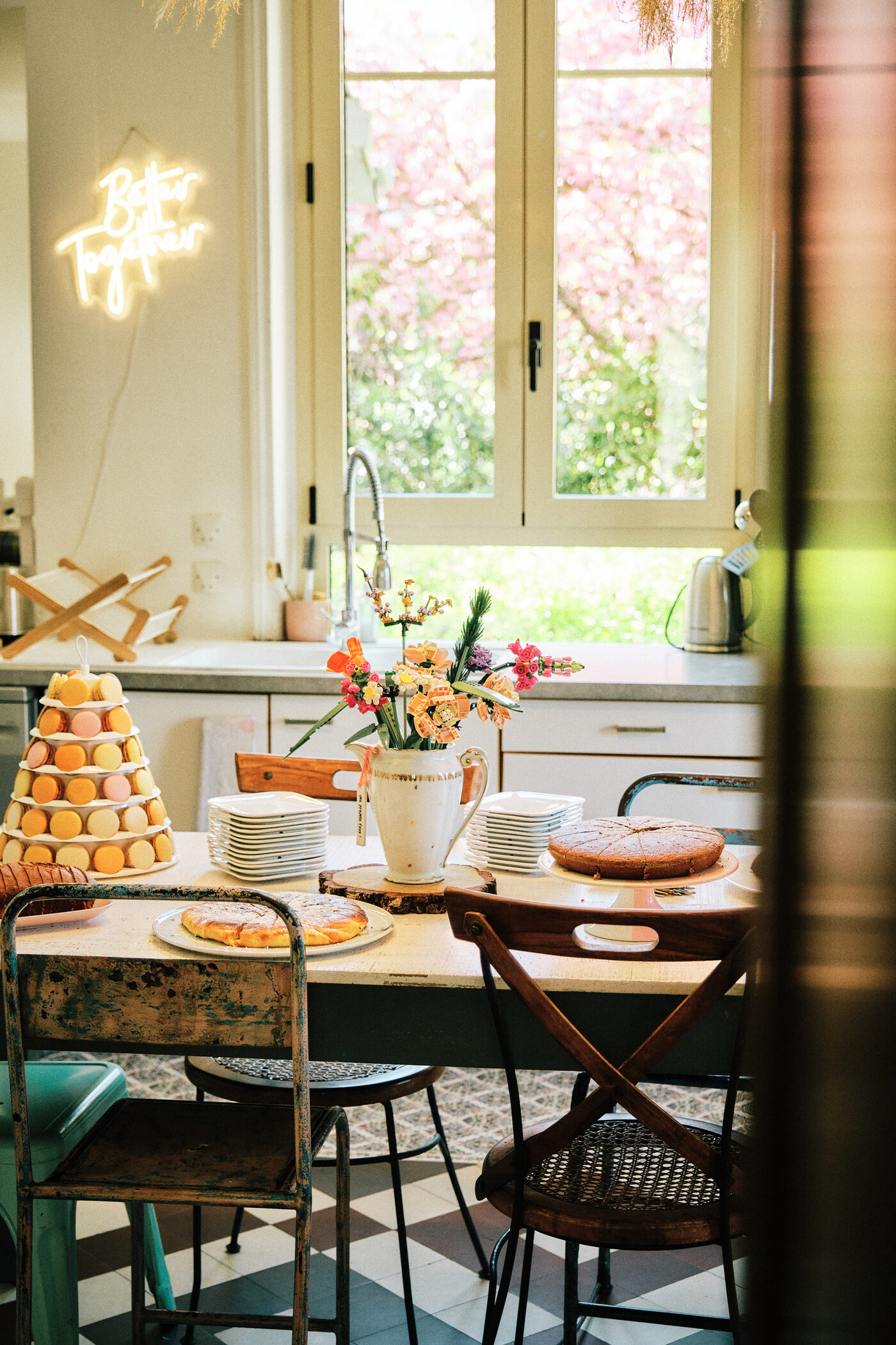 La table de la cuisine de la Villa Nymphea : une pyramide de macarons, deux tartes, des piles d’assiettes et un pichet de fleurs, devant la fenêtre ouverte sur le cerisier en fleur