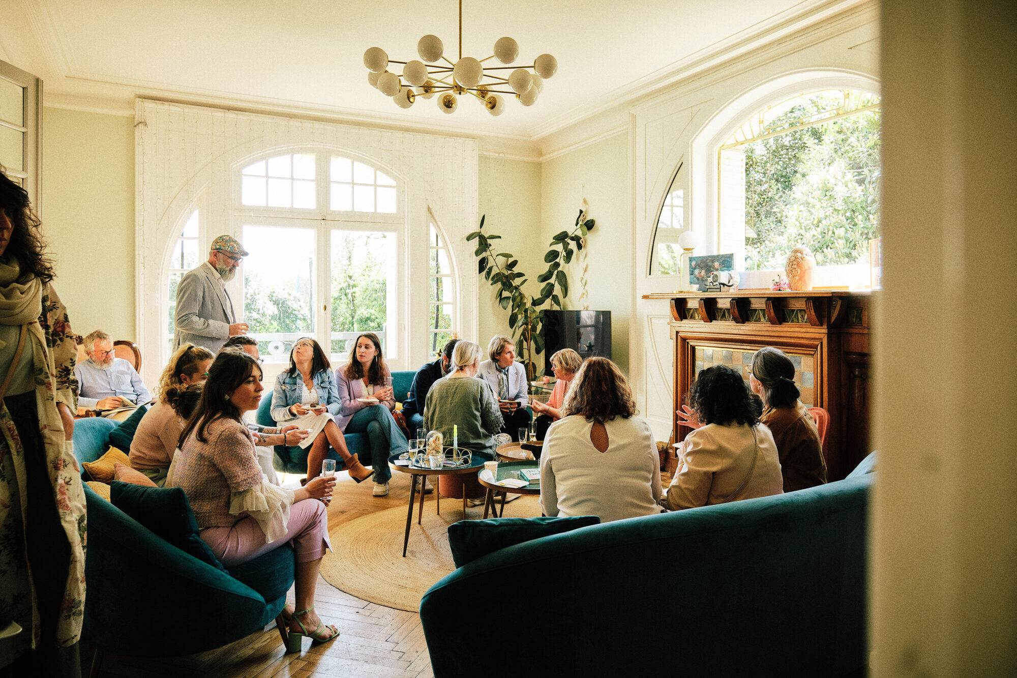 Le salon de la Villa Nymphea : une vingtaine de personnes assises dans les fauteuils de velours bleu, un verre à la main, devant les grandes fenêtres en arc et la cheminée de faïence
